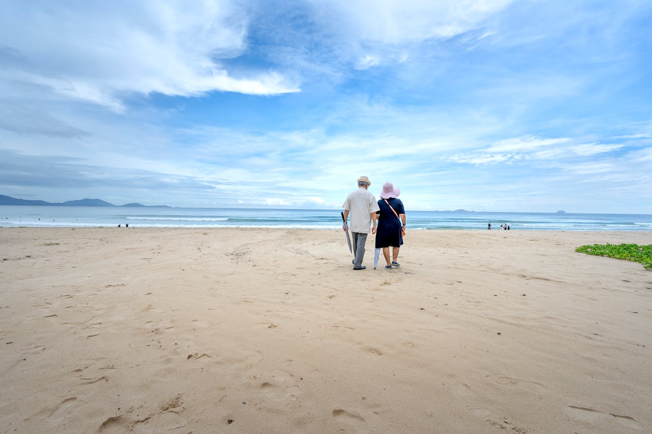A senior couple walking on a serene sandy beach under a beautiful cloudy sky.